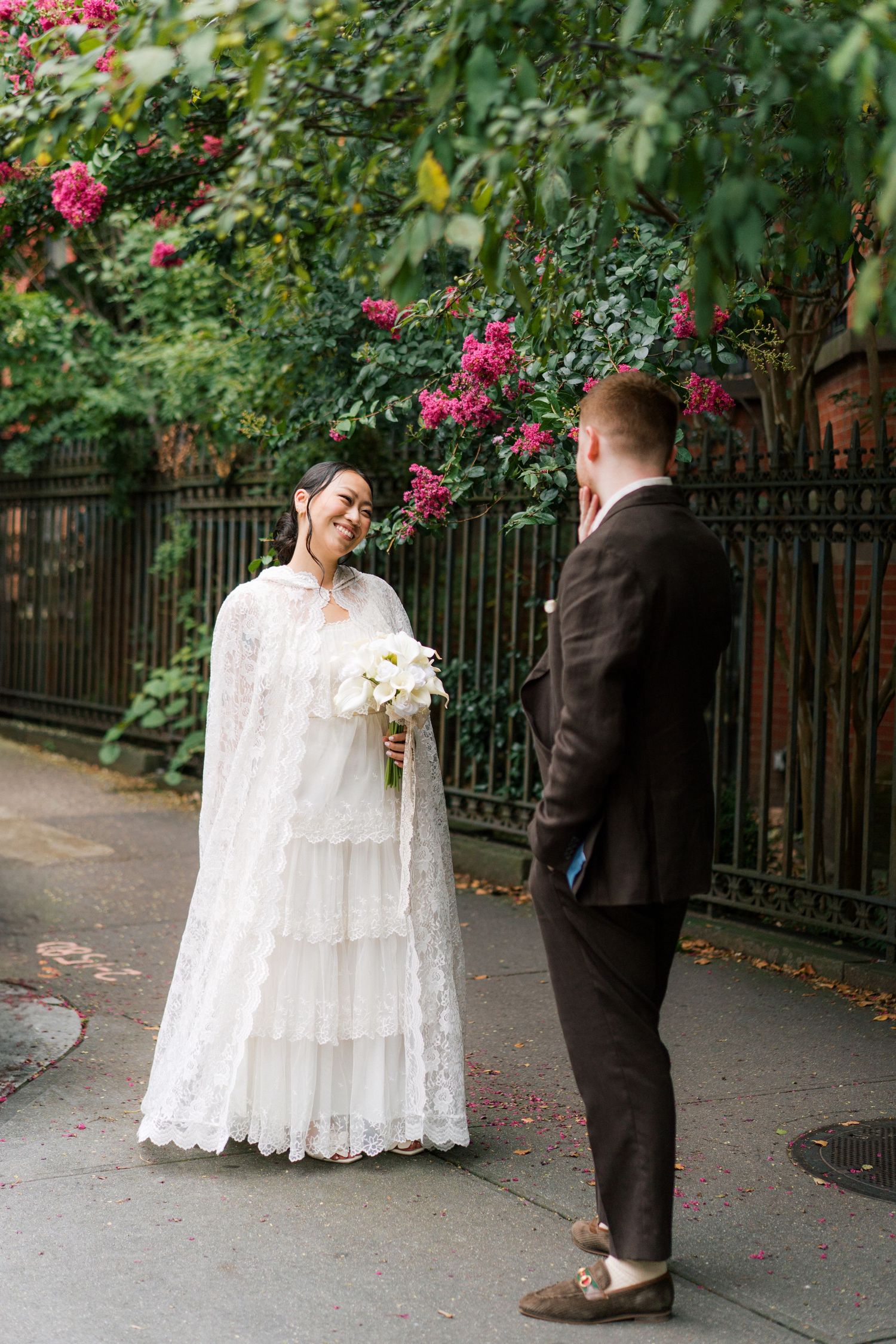Wedding couple poses in garden setting with blooming pink flowers and iron fence.