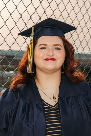 Graduate in navy cap and gown wearing striped shirt smiles in front of chain link fence background.