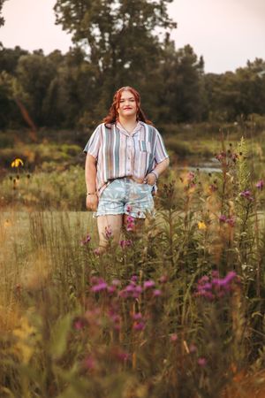 A person stands in a wildflower meadow at sunset wearing a striped shirt and floral shorts among purple and yellow blooms.