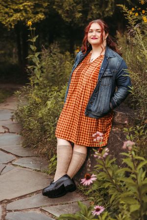 Person in orange gingham top and denim jacket poses near pink flowers on stone path in garden setting.