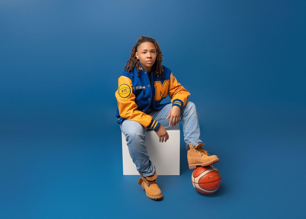 Shank Henry Media photo of basketball player in yellow and blue uniform sits on white block against blue backdrop with orange basketball.