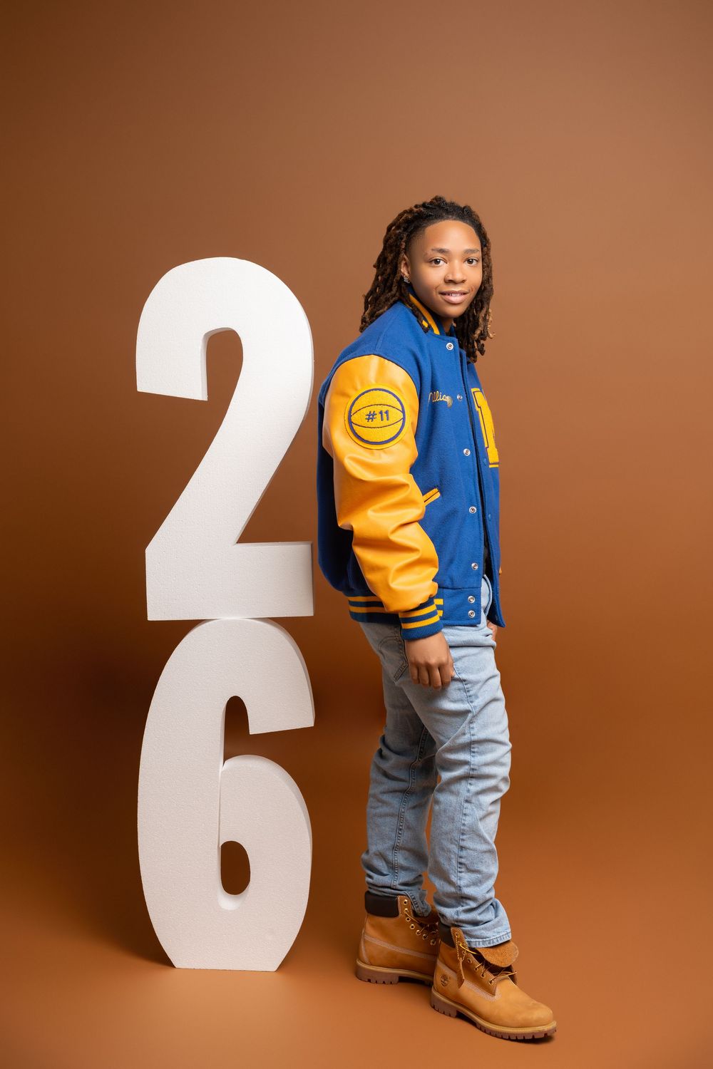 Shank Henry Media senior photo with stylish varsity jacket and jeans outfit with large white decorative numbers against brown backdrop.
