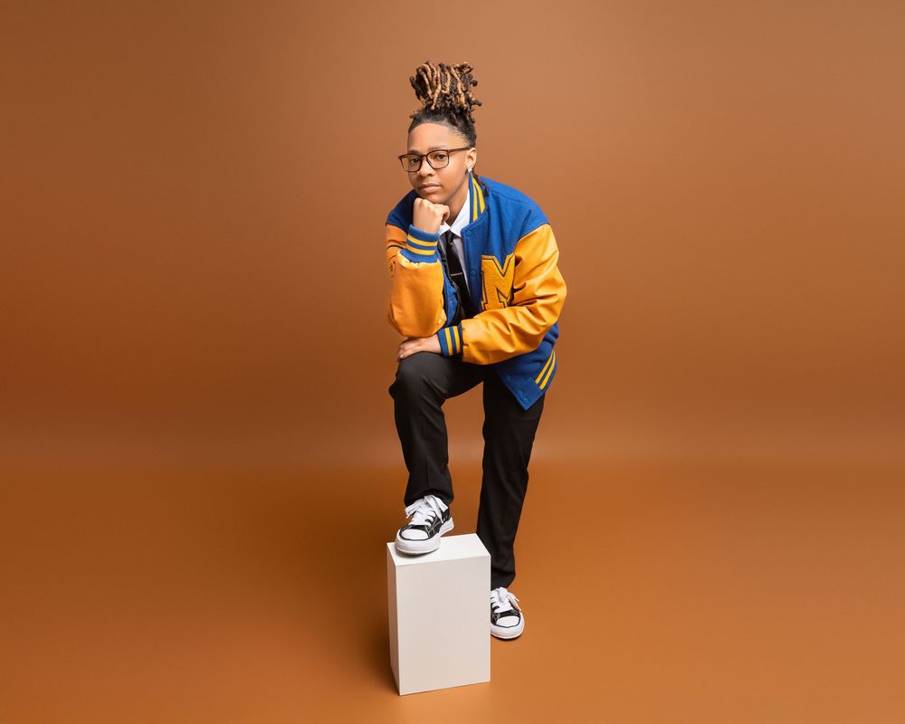 High school senior in varsity jacket and black pants sits on white cube against orange background while looking at phone.