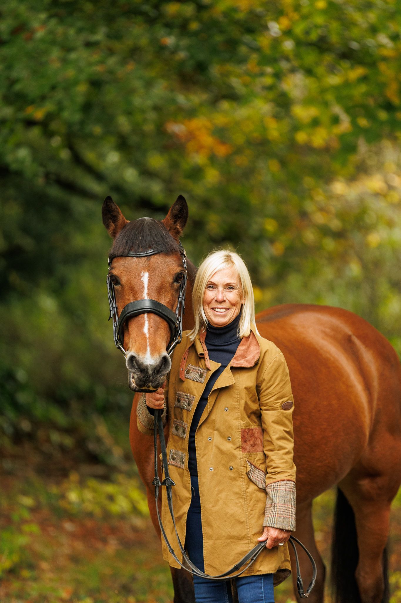 A brown horse and person in a tan jacket stand together in a wooded autumn setting.