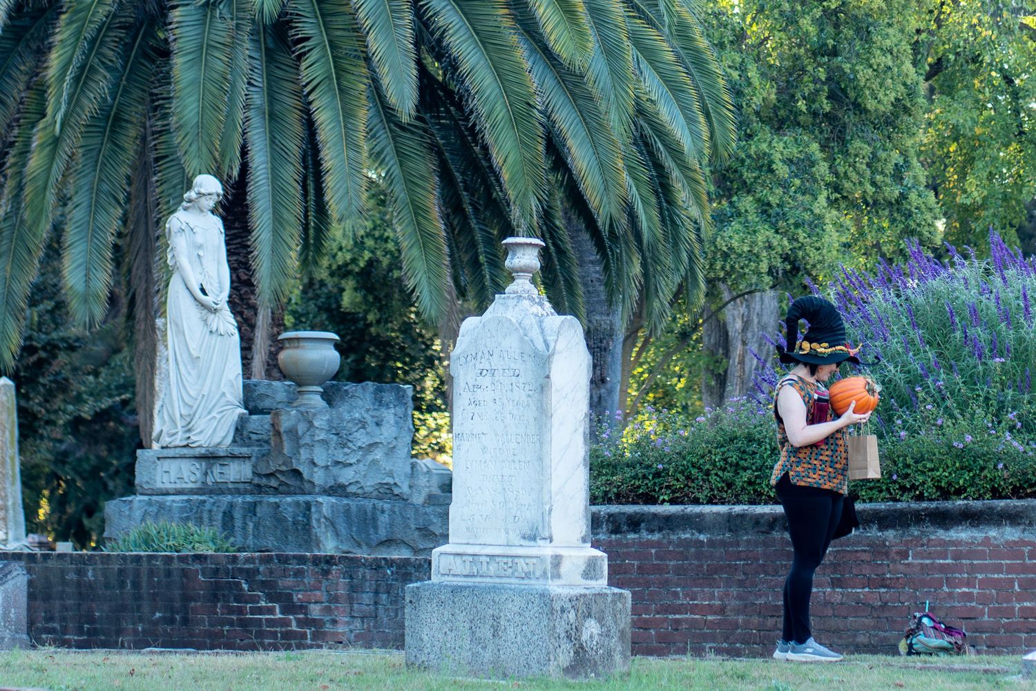A person walks past a row of white statues in a lush garden setting with palm trees and flowering plants.
