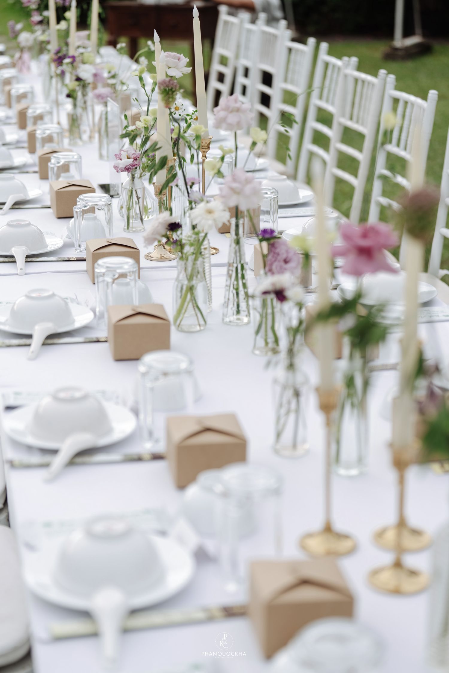 Elegant outdoor wedding reception table with white linens, floral arrangements, candles, and kraft paper favor boxes.