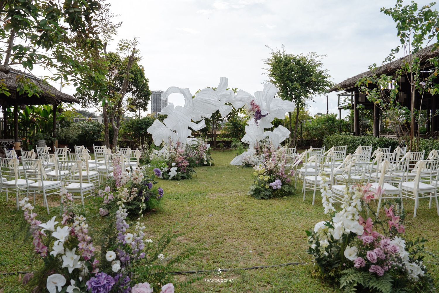 Outdoor garden wedding ceremony setup with white chairs, floral aisle decor, and a white orchid arch.