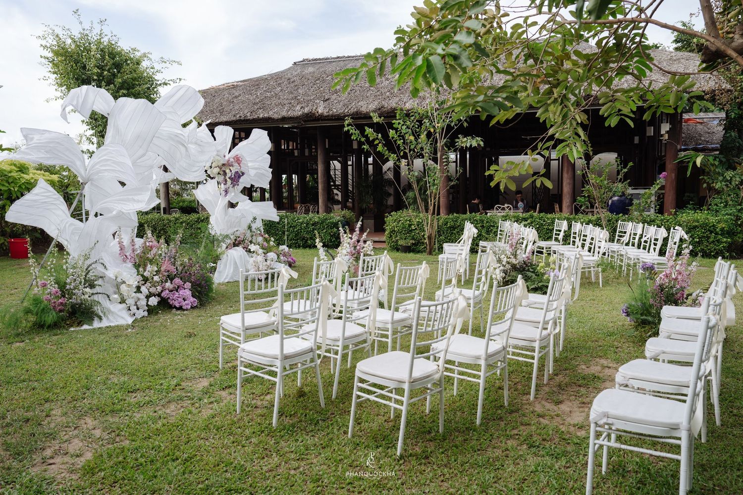 White Chiavari chairs arranged on green lawn with large white floral arch for an outdoor wedding ceremony.