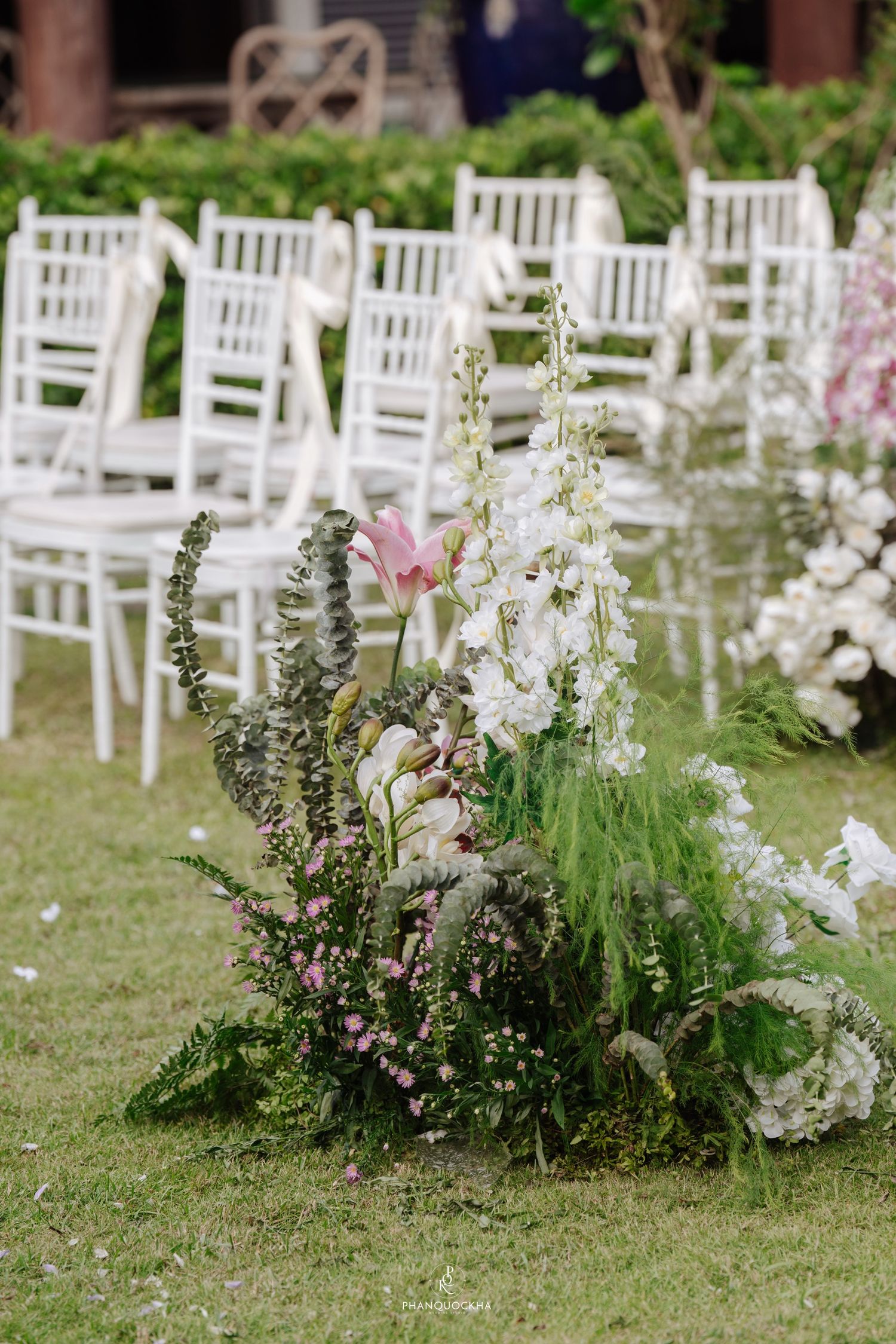 Lush garden wedding aisle floral arrangement with white delphiniums, pink lilies, and greenery beside white chairs.