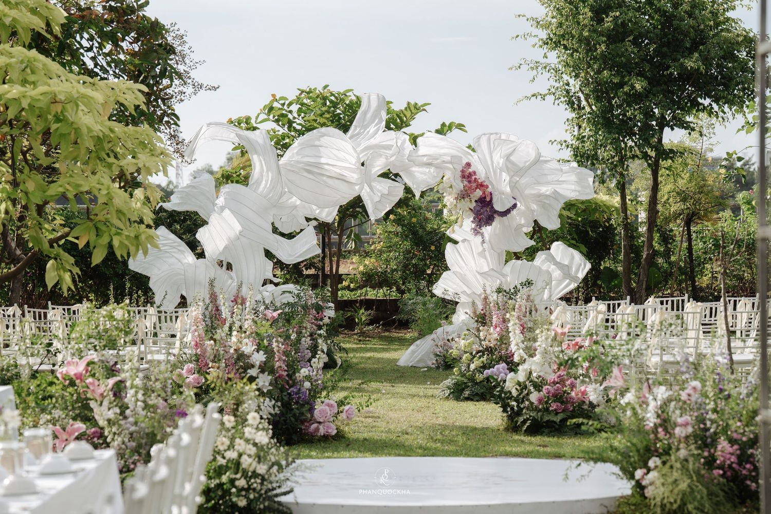Stunning outdoor garden wedding ceremony with white sculptural arch, floral aisle decor, and white Chiavari chairs.