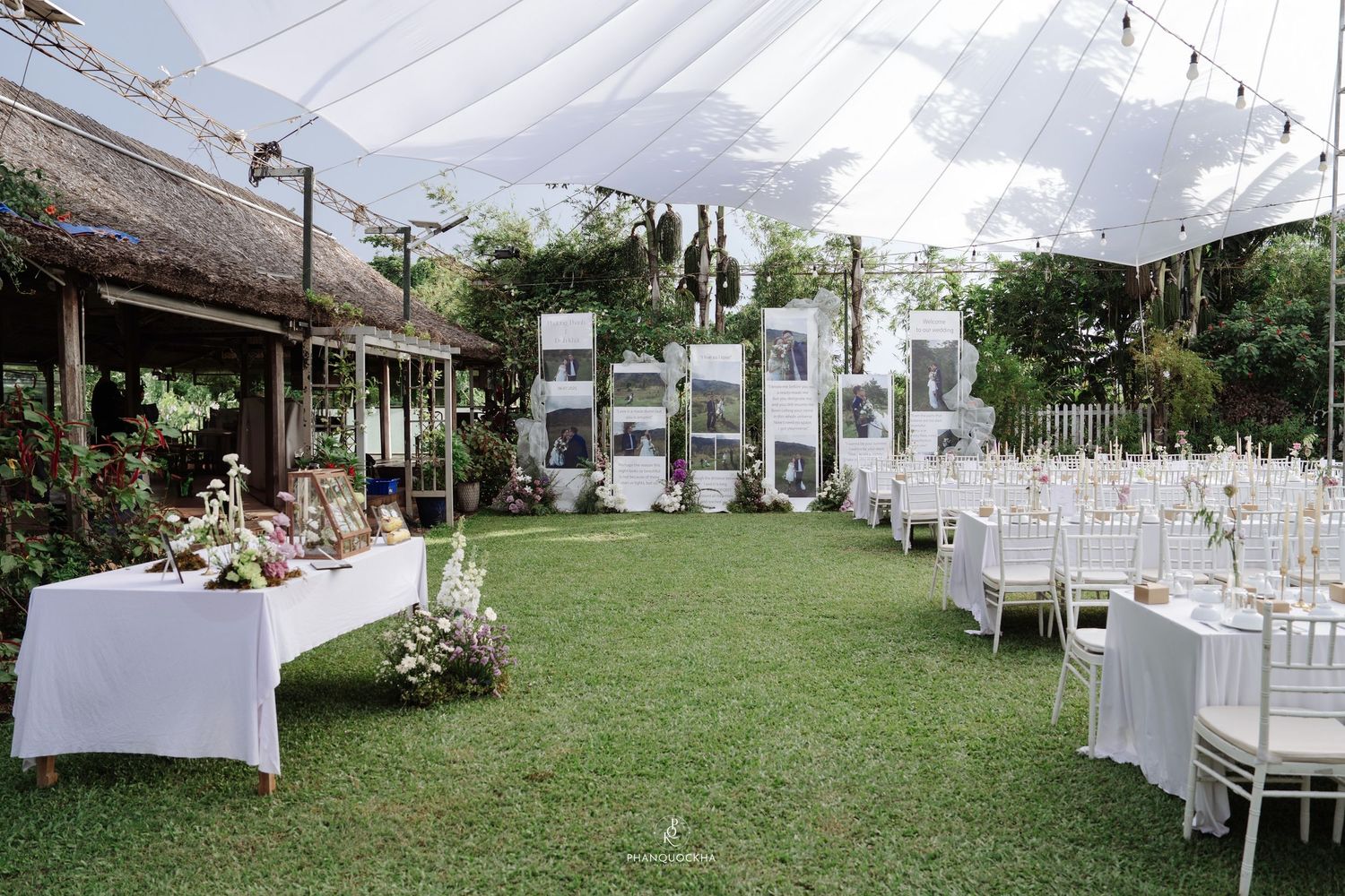 Outdoor wedding reception venue with white draped ceiling, photo display wall, and white-clothed dining tables.