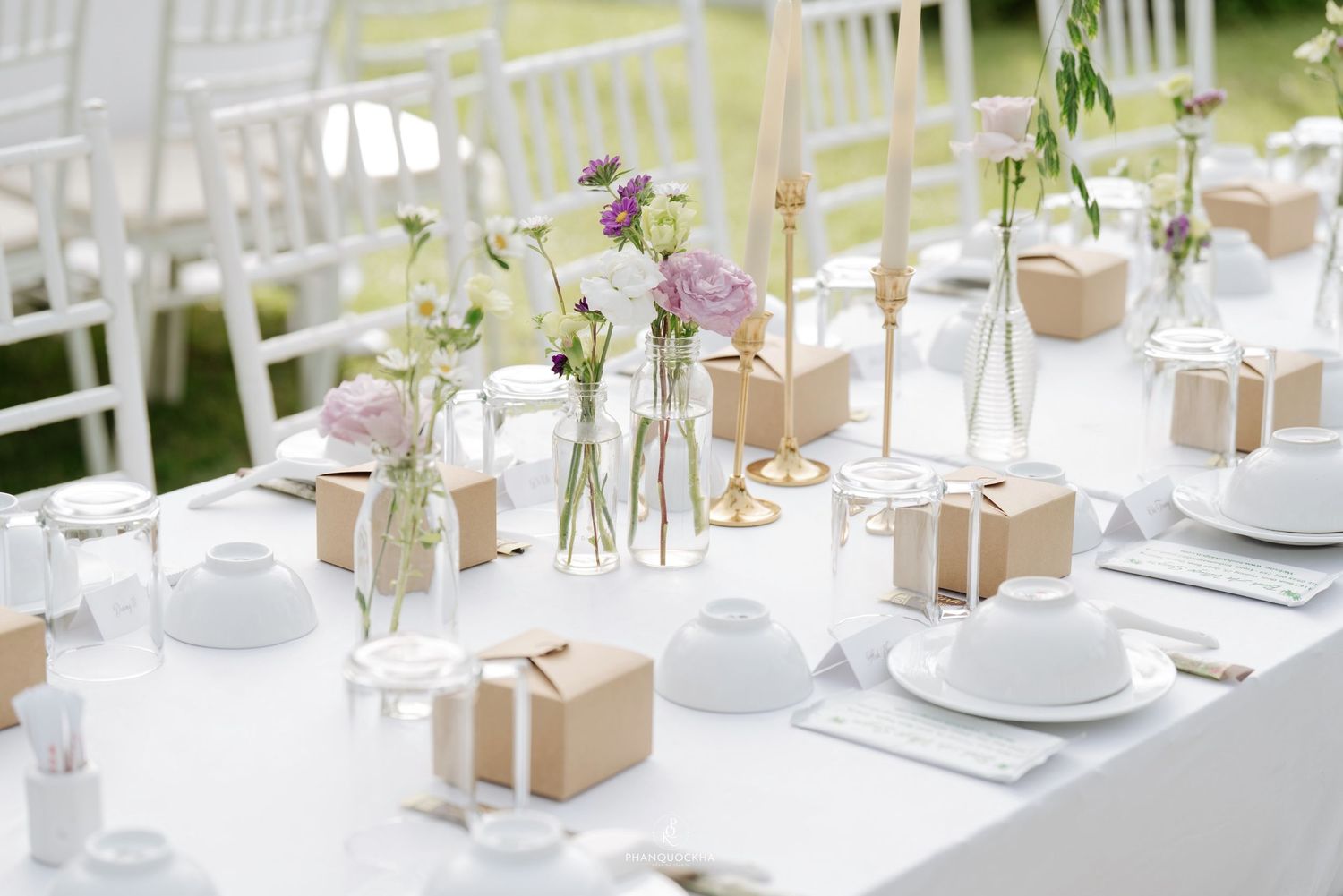 Elegant wedding reception table with white settings, kraft favor boxes, gold candle holders, and purple florals.