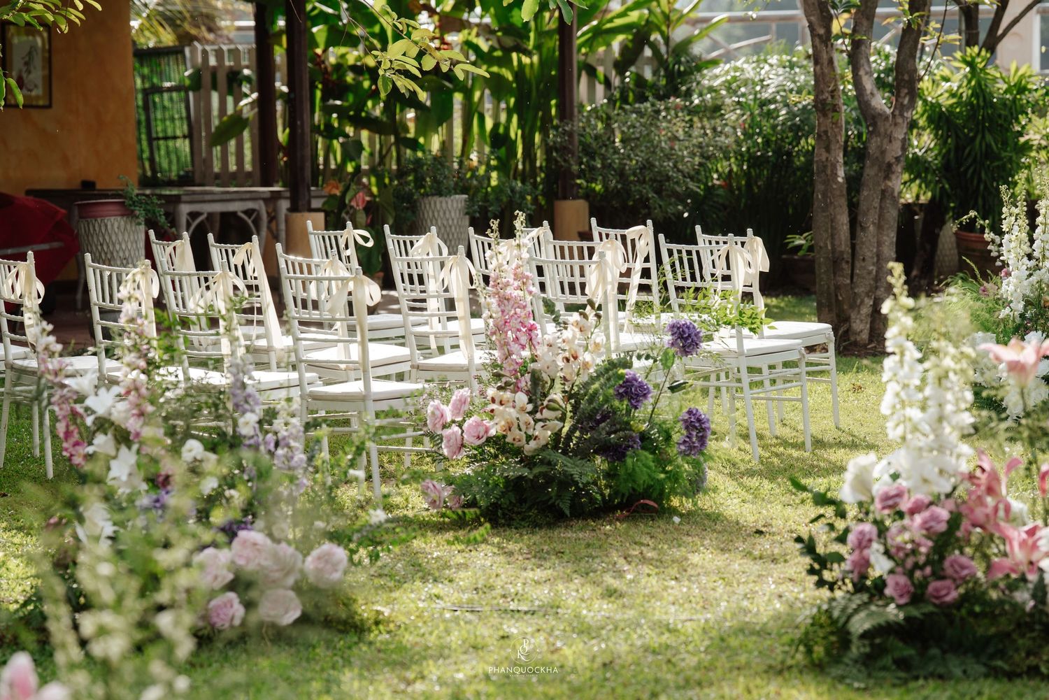 Garden wedding ceremony with white Chiavari chairs, colorful floral aisle arrangements of pink and purple blooms.