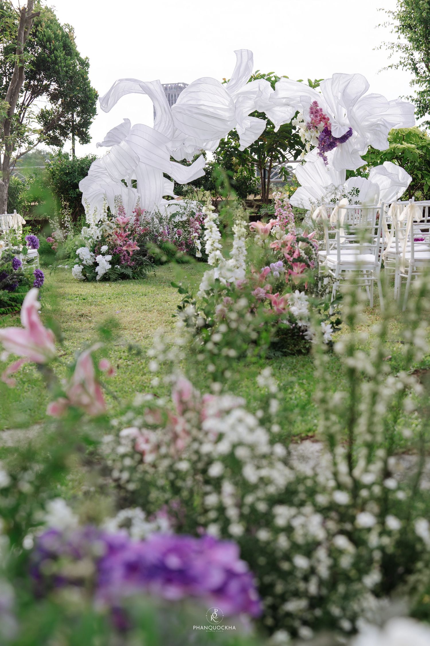 Elegant outdoor wedding ceremony aisle lined with pink and white flowers, featuring large white fabric installations as backdrop.