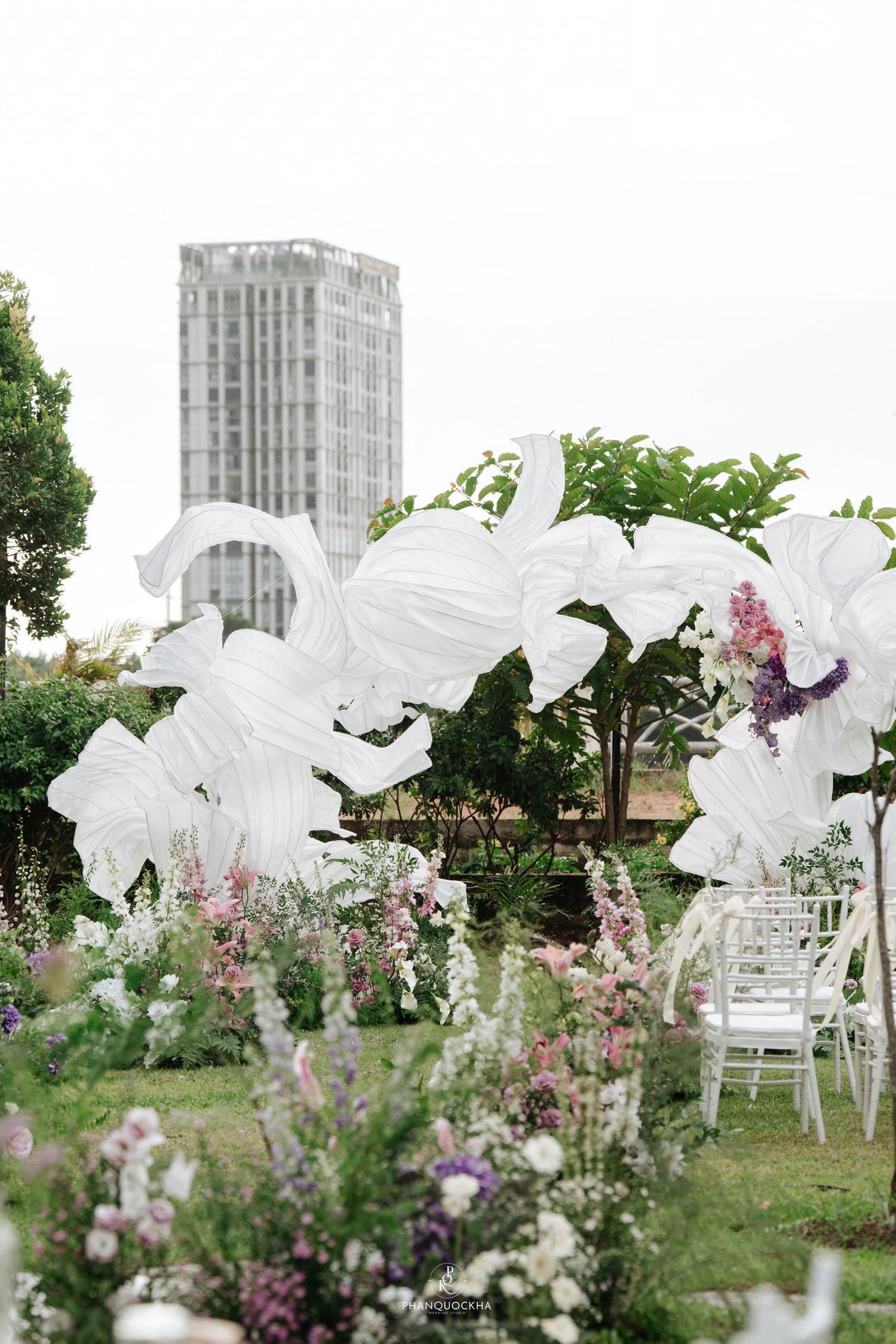 Outdoor garden wedding venue with oversized white fabric floral sculptures and pink blooms, city building visible in background.