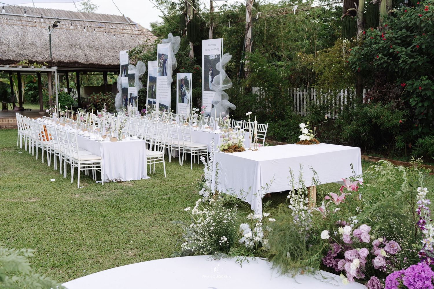 Outdoor wedding reception setup with long white-draped tables, chiavari chairs, and black-and-white photo displays on green lawn.