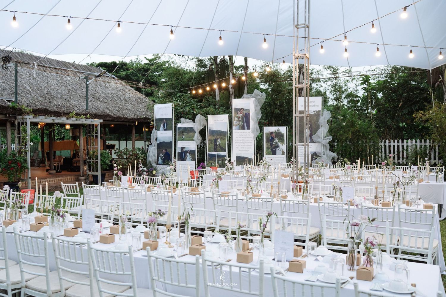 Elegant outdoor wedding reception under string lights with long white tables, chiavari chairs, and decorative photo displays.