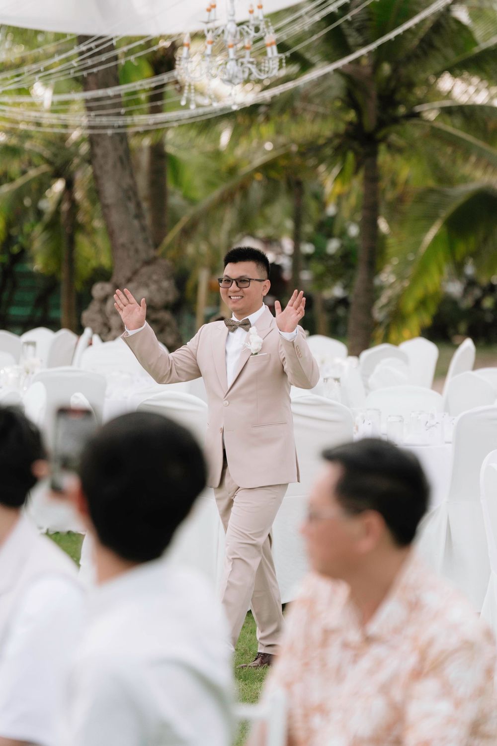 Groom in beige suit with bow tie raises hands joyfully at outdoor garden wedding ceremony.