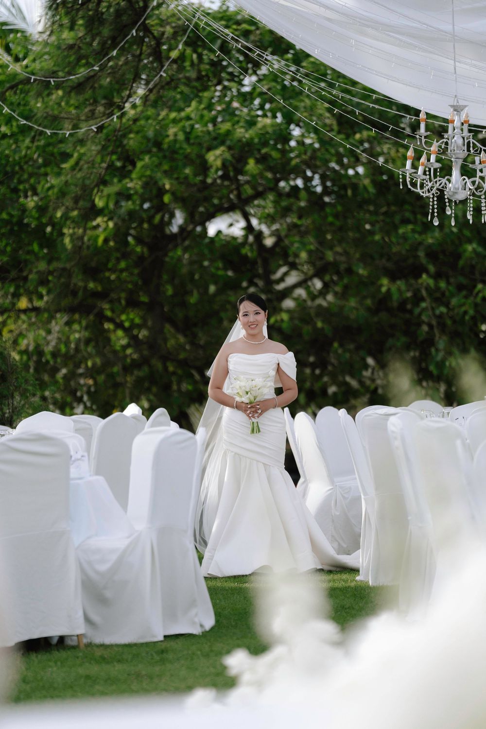 Bride in white off-shoulder gown and veil walks down aisle at elegant outdoor garden wedding ceremony.