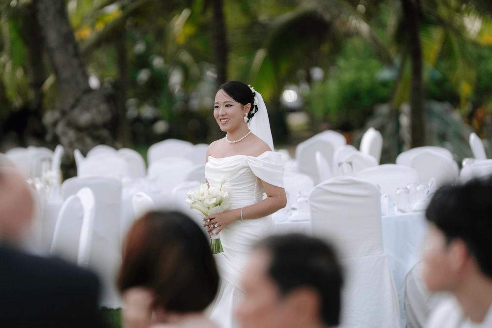 Radiant Asian bride in white off-shoulder dress smiles while holding white bouquet at outdoor wedding.