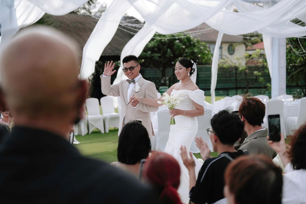 Groom waves to guests while standing beside bride at elegant outdoor garden wedding ceremony.