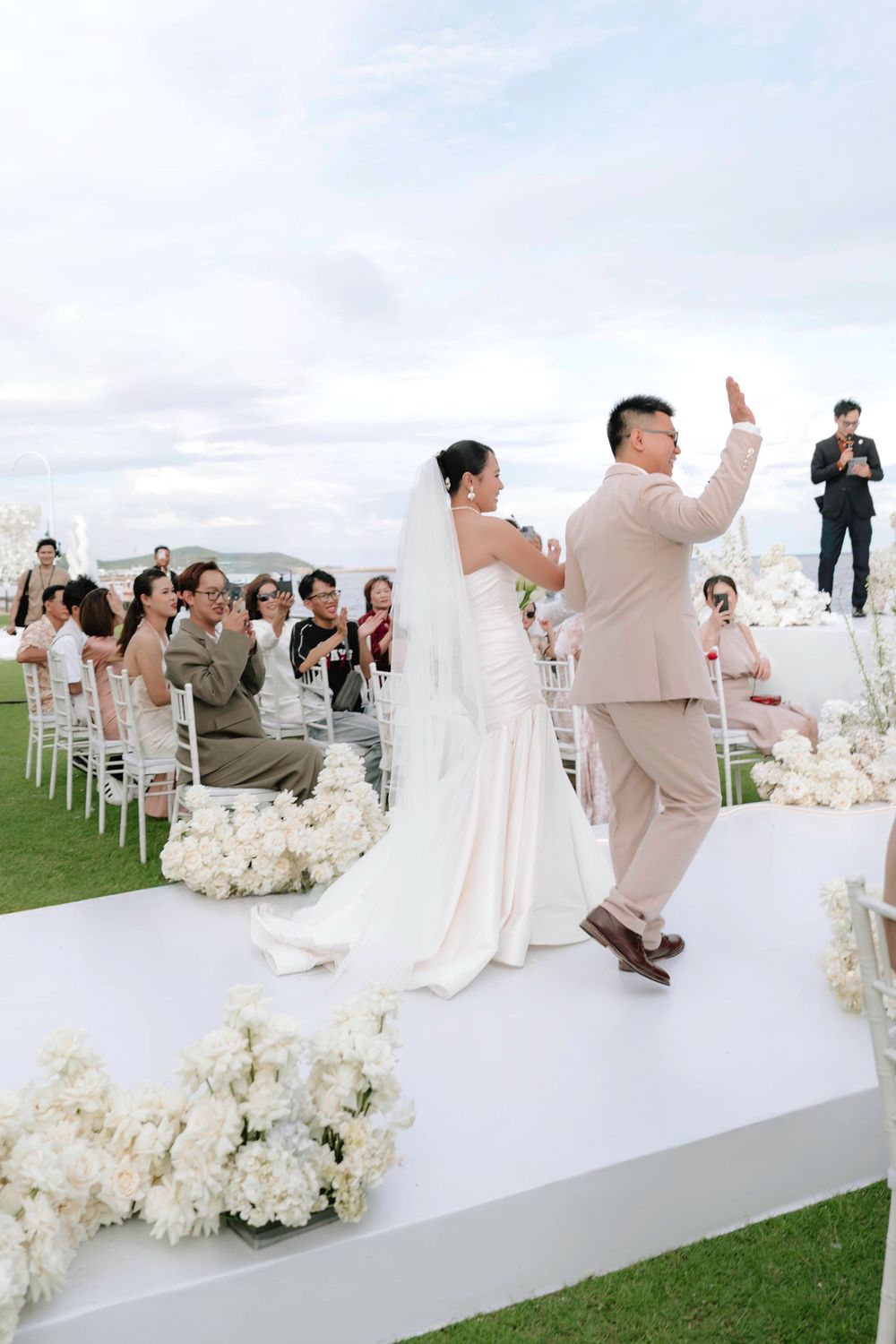 Newlywed couple walks down white aisle as guests applaud at elegant outdoor wedding ceremony.