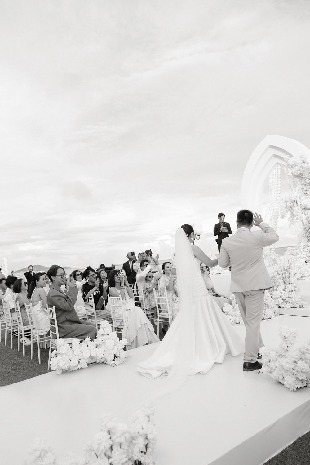 Black and white photo of bride and groom at outdoor wedding ceremony with seated guests applauding.