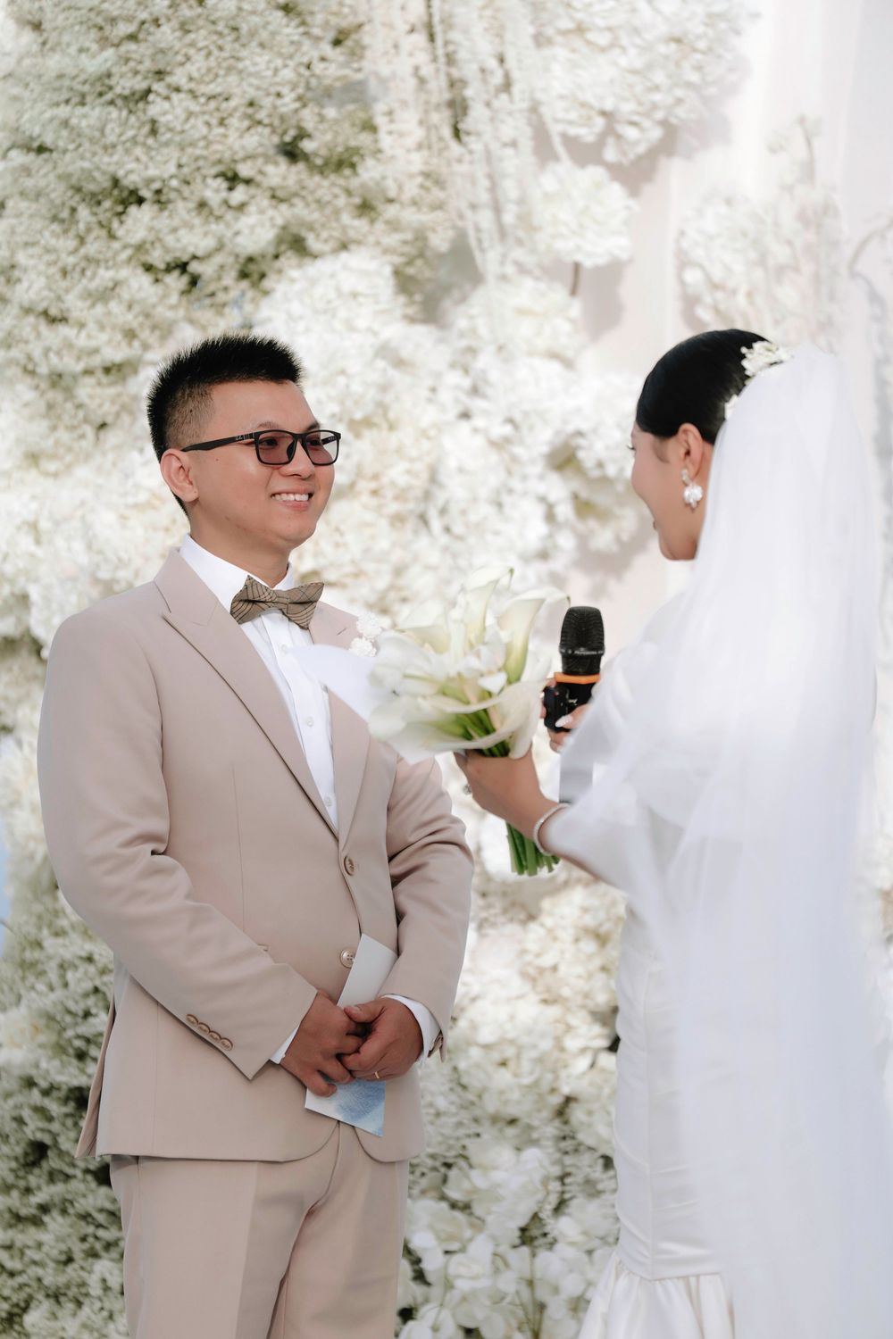 Groom in beige suit smiling at bride during outdoor wedding vow exchange.