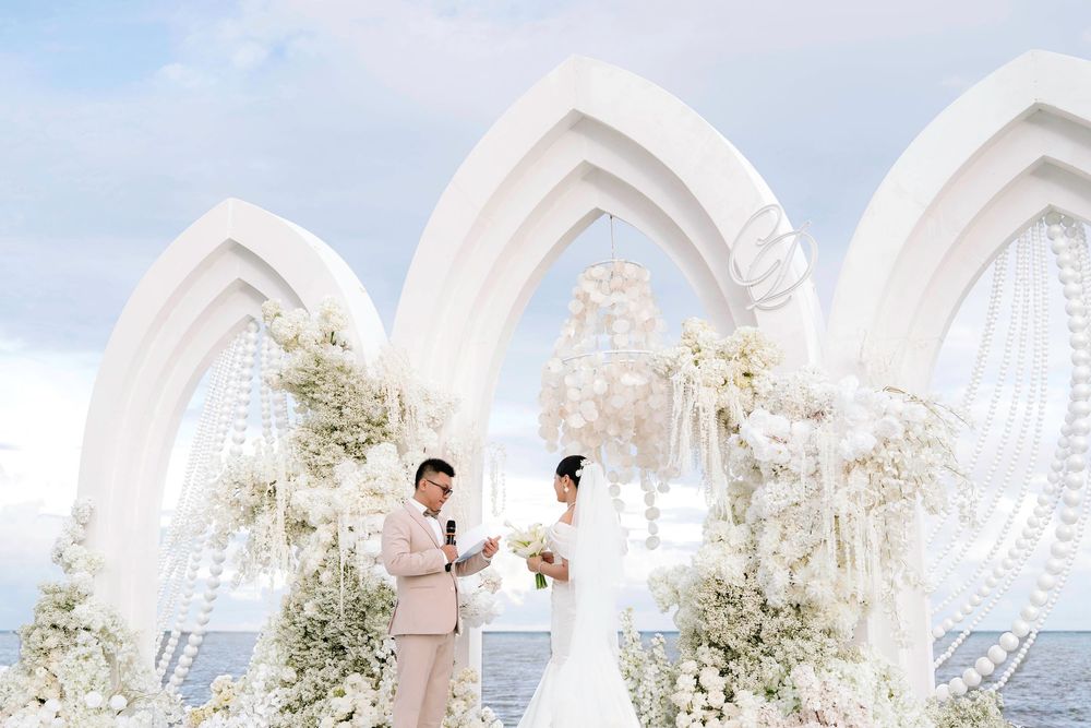 Groom reading vows to bride under white floral arches at ocean wedding.