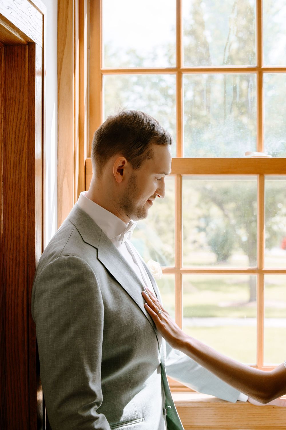 Groom in gray suit looks pensively out large wooden-framed windows in bright natural light.