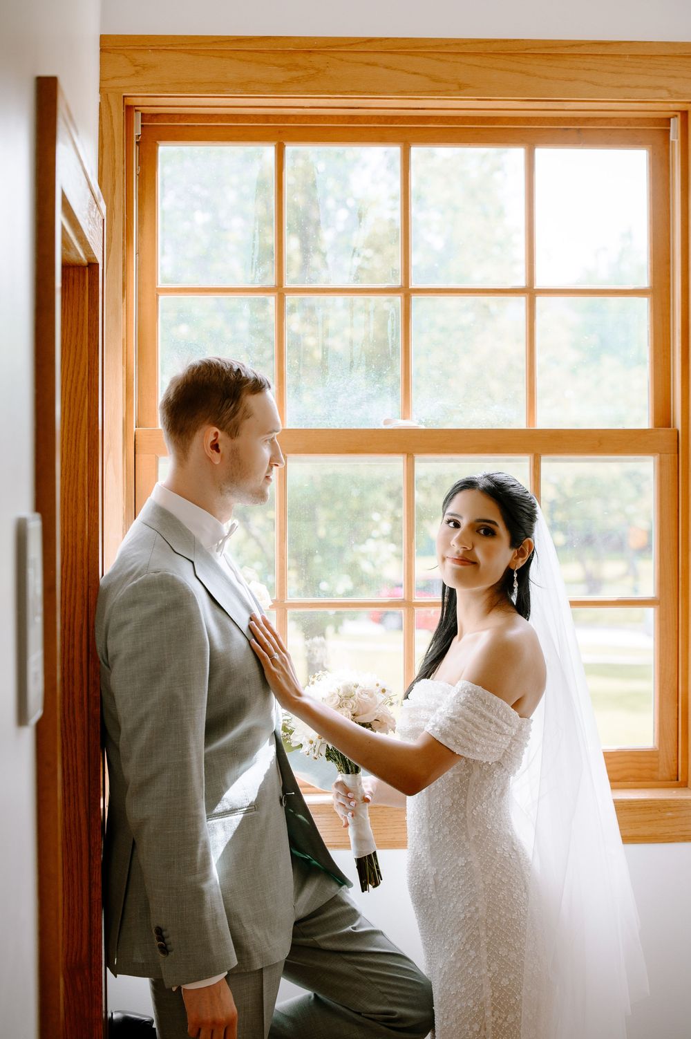 A couple in wedding attire shares a playful moment near large windows.