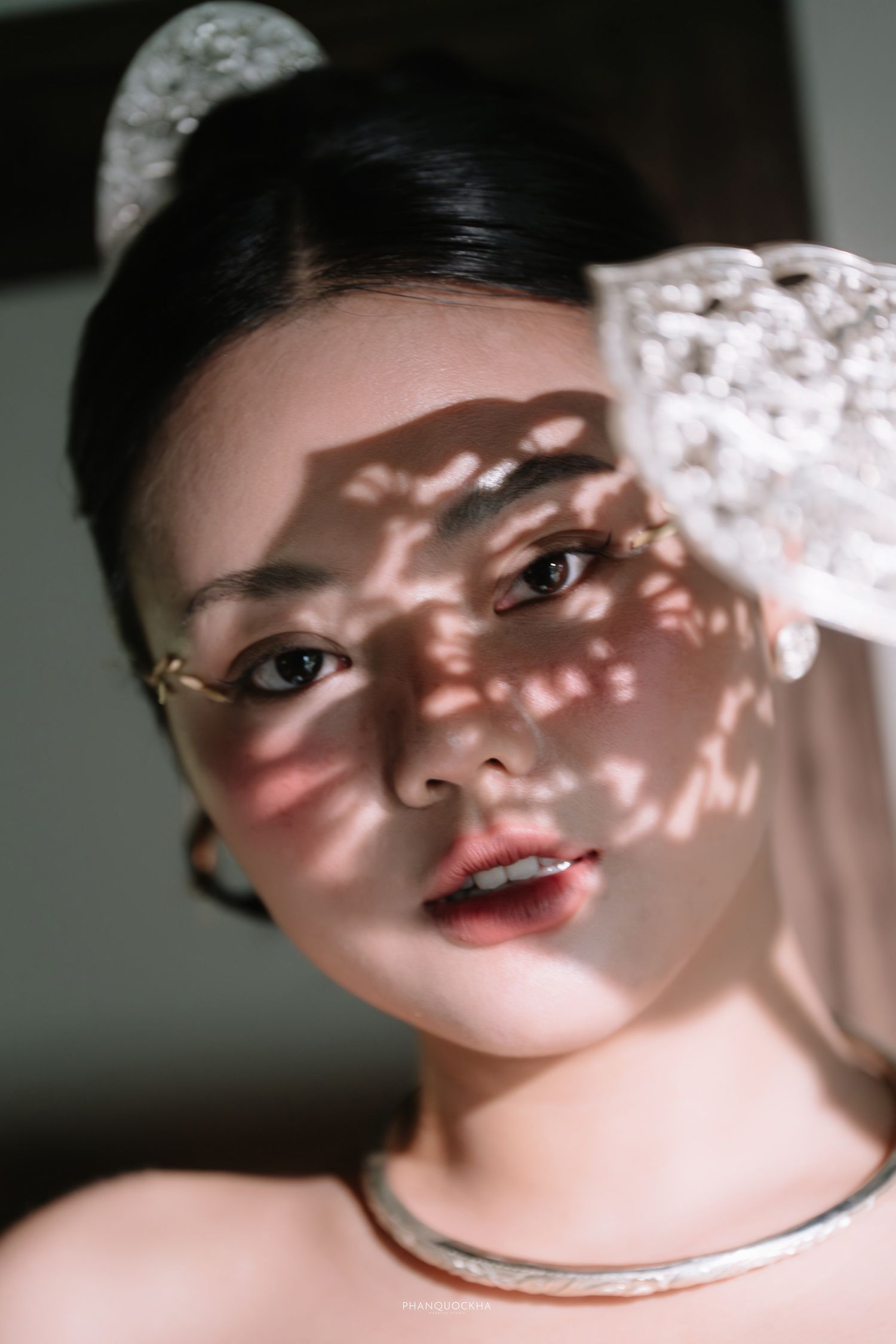 Young Asian woman with elegant updo, lace hair bow, rimless glasses, and silver necklace, with sunlight shadow patterns on face.
