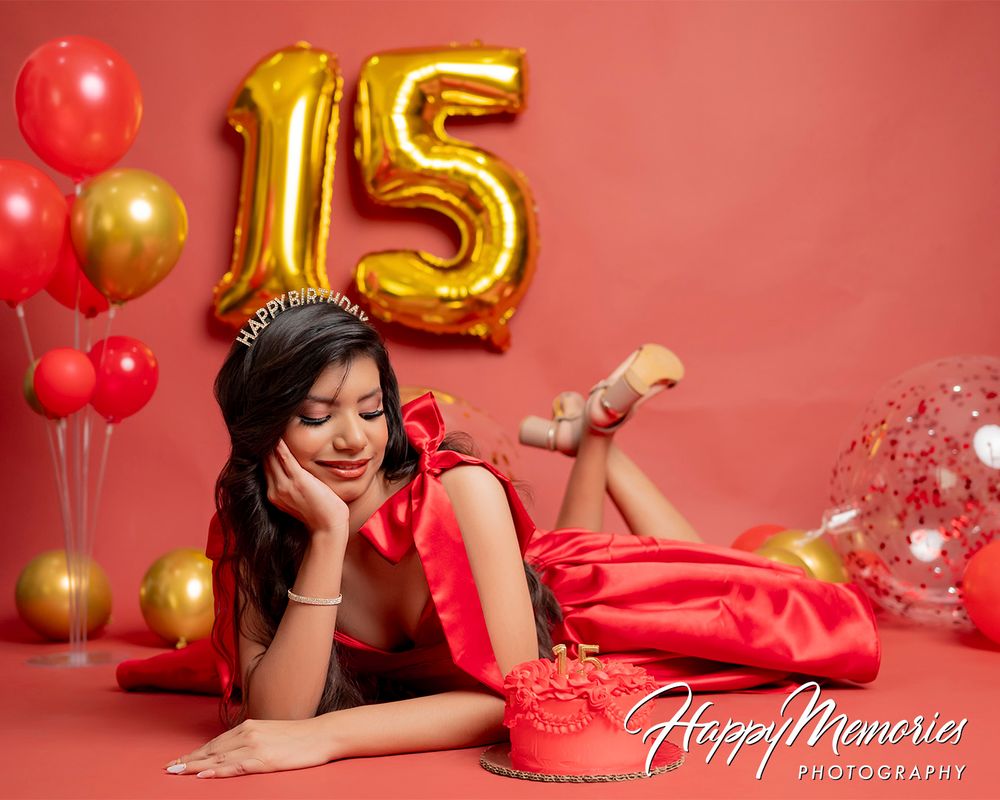 Birthday celebration photoshoot with red dress and metallic number 15 balloons on coral backdrop.