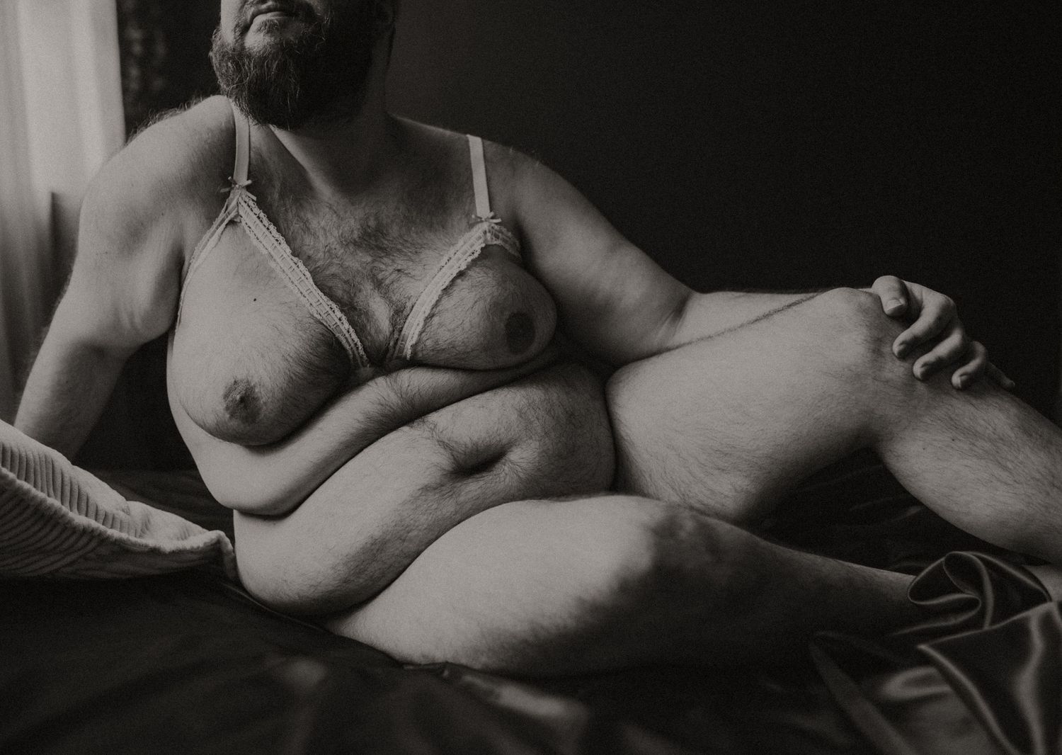 Plus size man in bed wearing white lace lingerie while relaxing comfortably on a bed surrounded by furry pillows and blankets.
