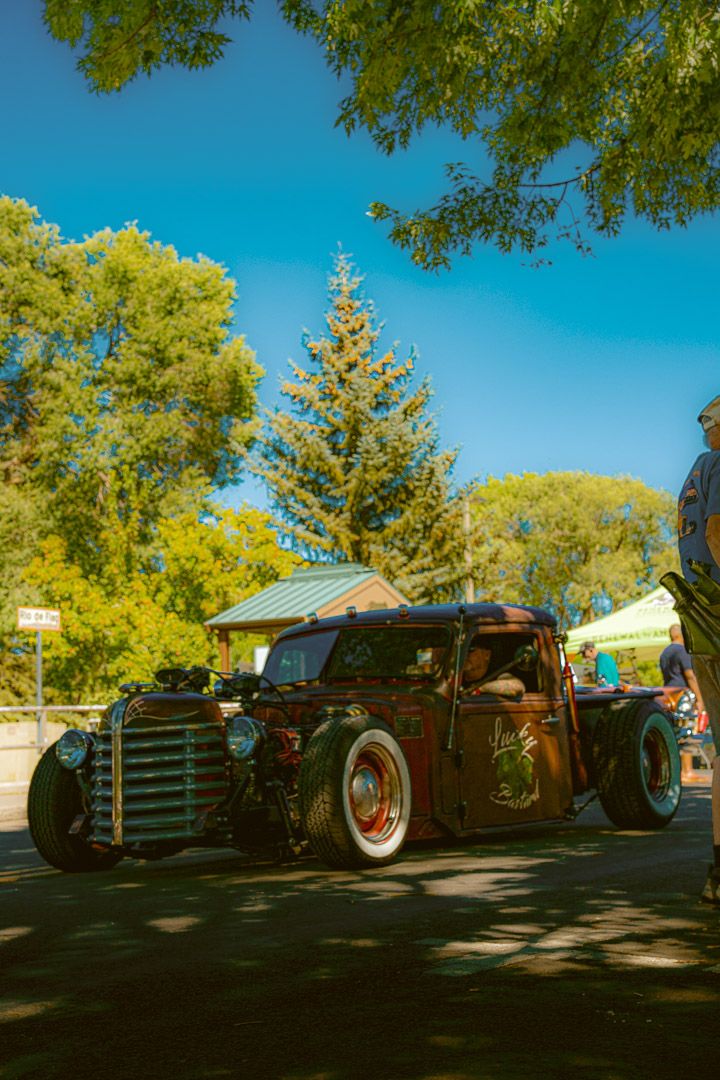 A row of classic vintage hot rods parked in the shade under green trees on a sunny day with blue skies.