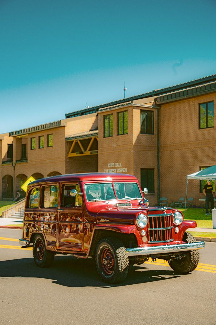 A row of vintage classic cars parked in front of a tan building on a sunny day, showcasing 1950s and 1960s automotive design.
