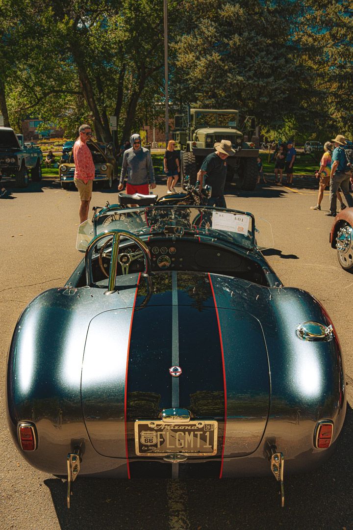 Classic silver Cobra roadster with racing stripes shown from multiple rear angles in a parking lot.