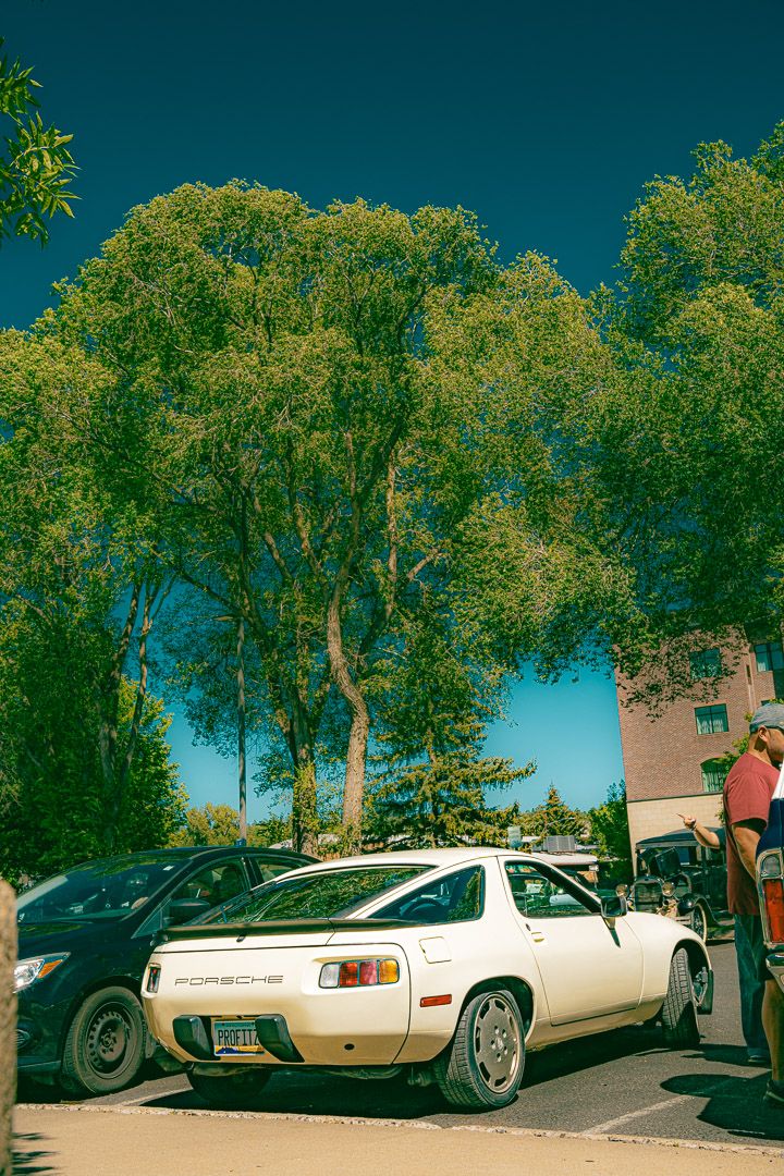 Row of classic cars parked in a lot with trees and buildings visible in the background.