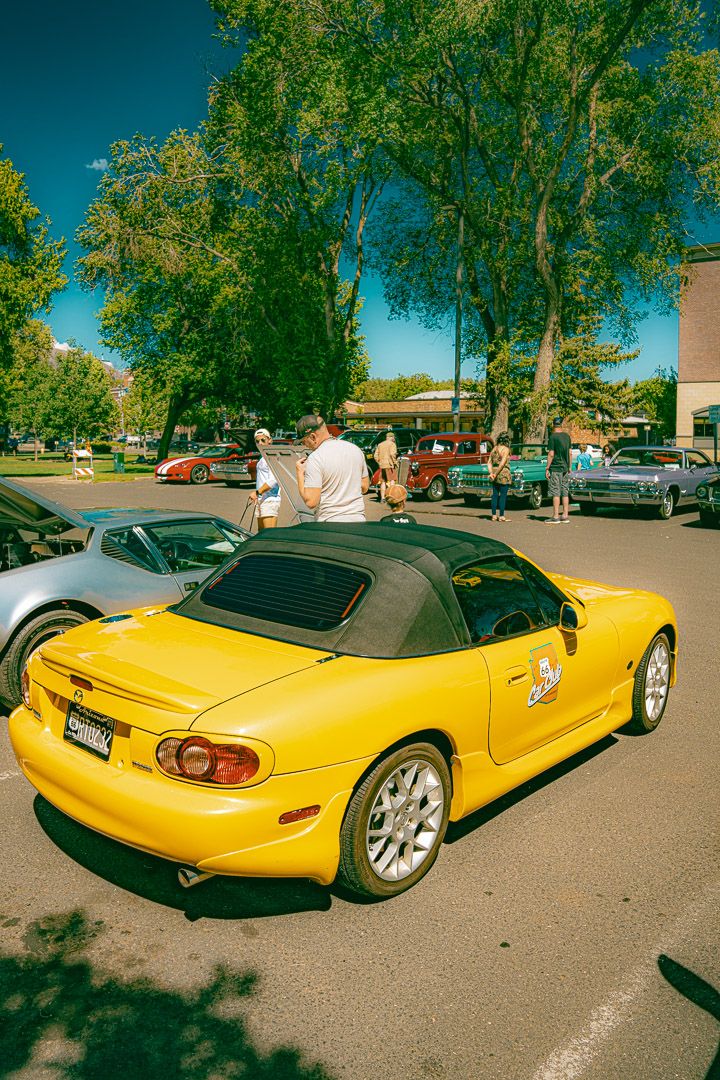 Yellow Miata convertible sports cars lined up in a sunny parking lot with trees in the background.