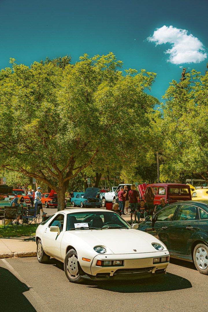 A sleek white vintage Porsche 928 sports car shown from multiple angles parked at a car show under green trees.
