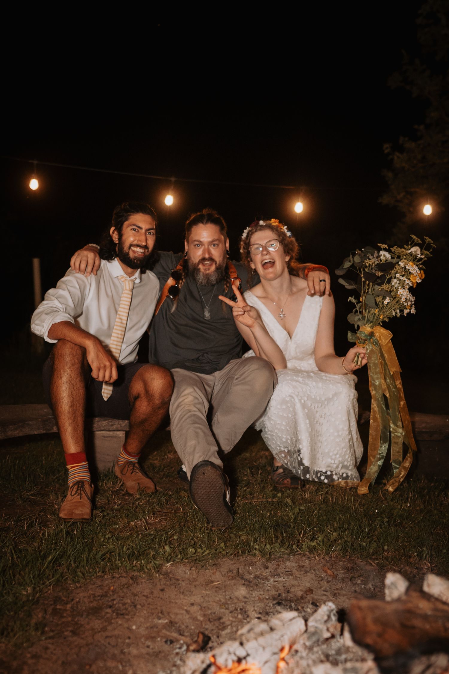 Three people laugh together while sitting outdoors at night with string lights hanging overhead and a campfire nearby.