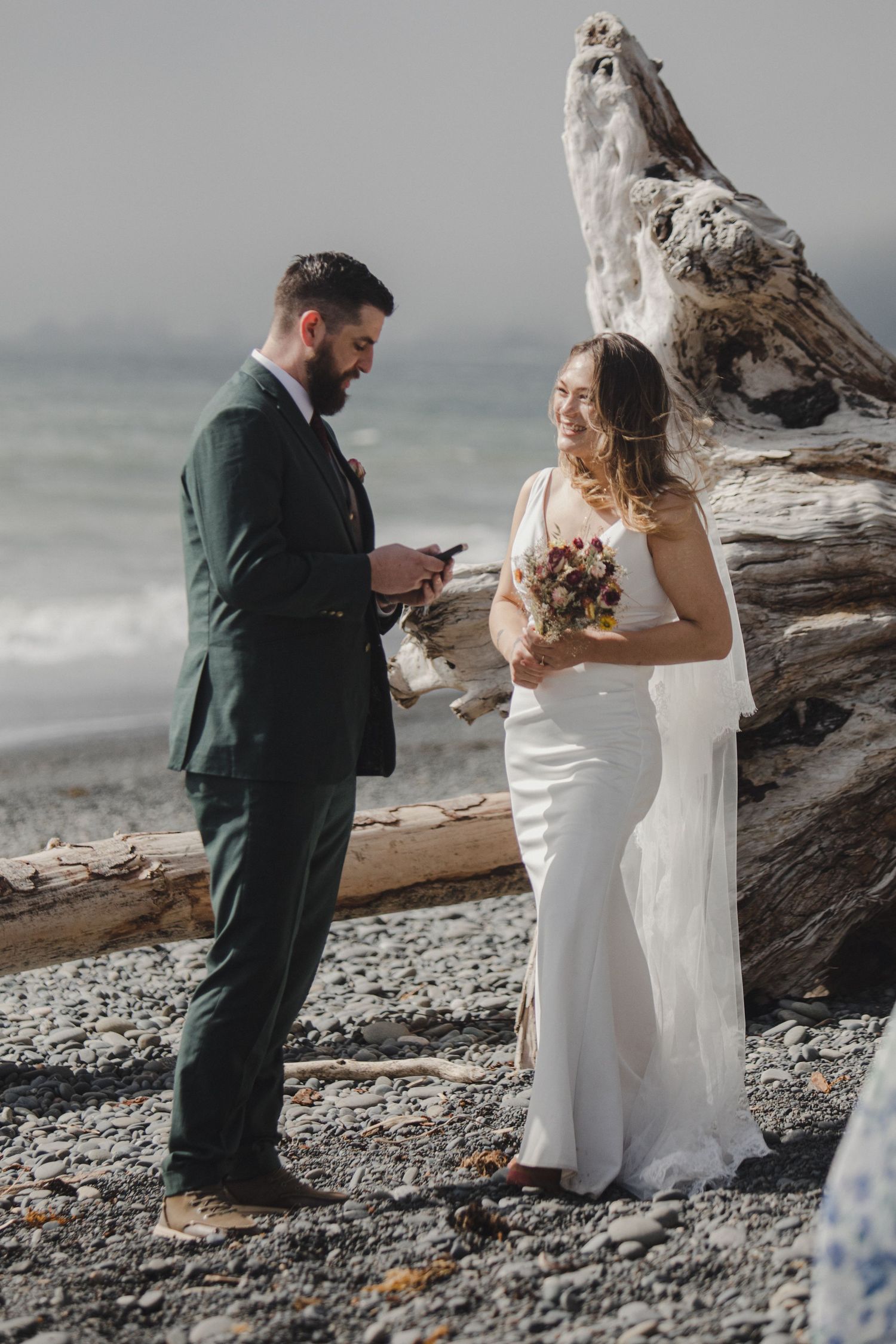 A beachside wedding ceremony takes place against a misty ocean backdrop with driftwood and rocky shore.