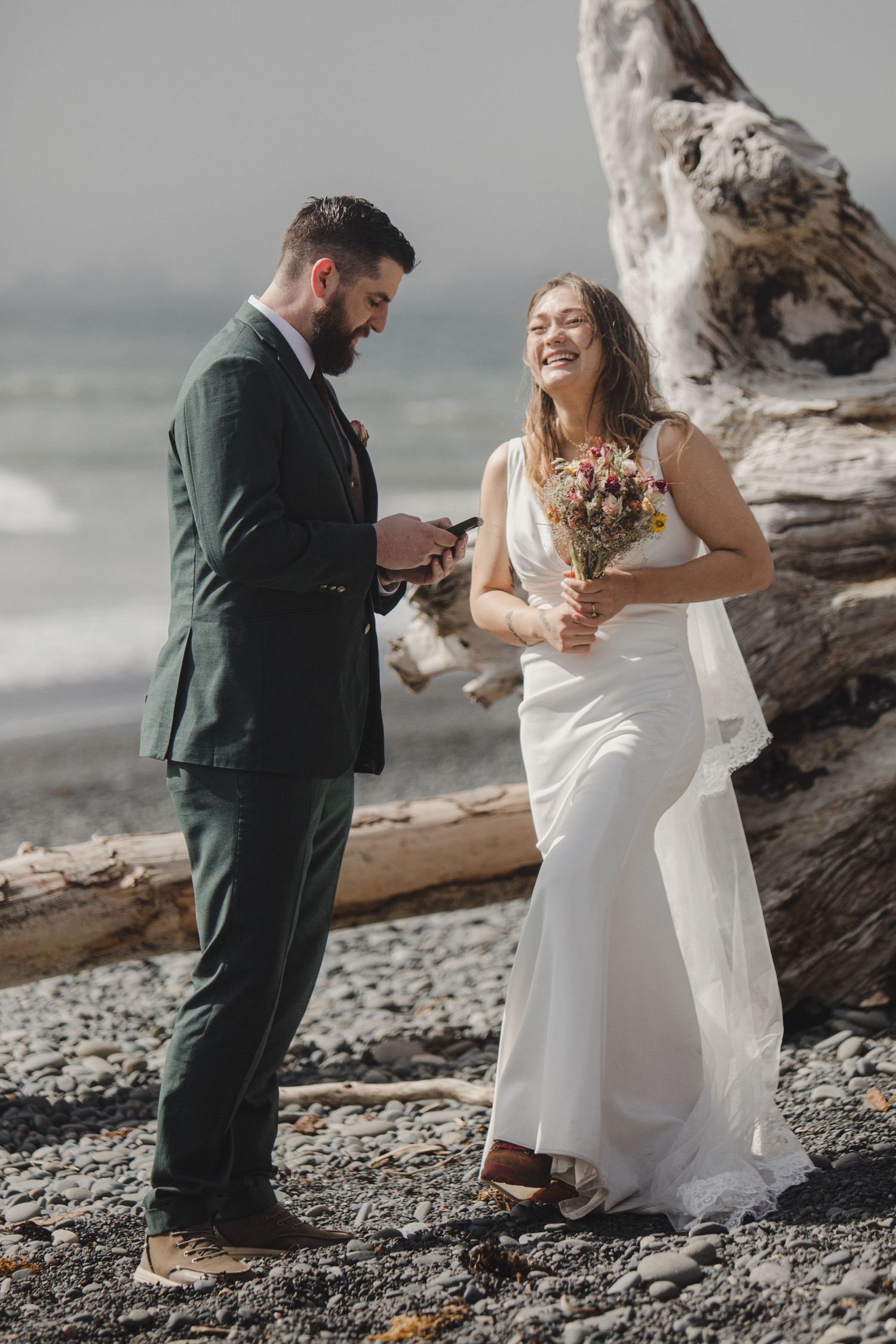 A couple sharing a joyful moment at their intimate beach wedding ceremony beside driftwood on a rocky shore.
