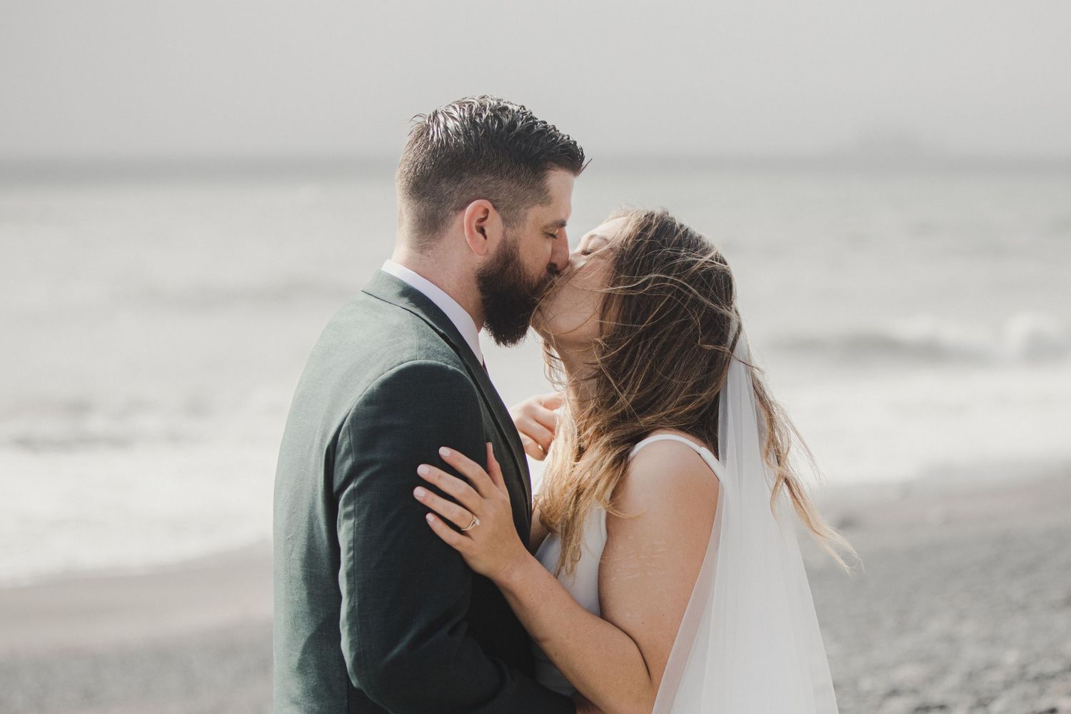 A romantic couple shares an intimate kiss on a misty beach while wearing wedding attire.