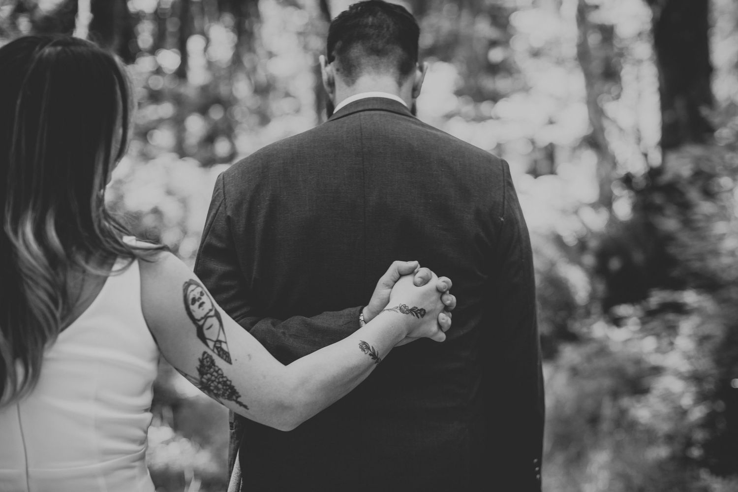Black and white photo showing a romantic couple holding hands while walking through a wooded forest setting.