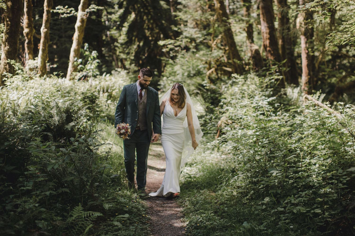 Couple walking hand in hand through a lush green forest path on their wedding day.