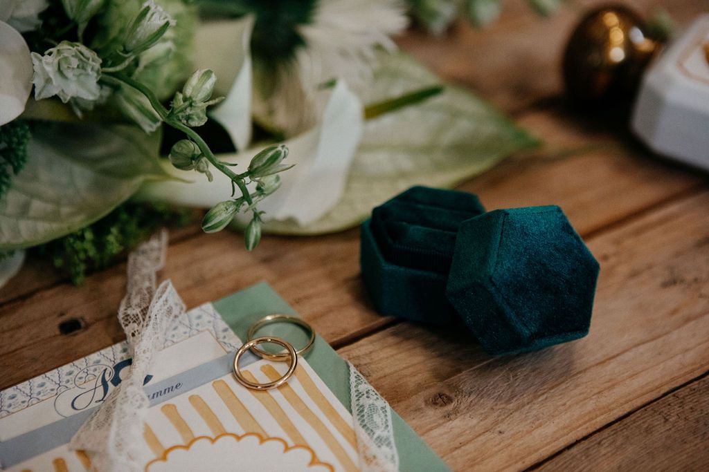 Wedding flat lay with gold rings, teal velvet ring box, bouquet, and invitation on wood.