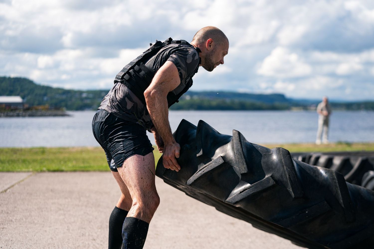 Person wearing black shorts and tank top exercises with a large tire by a lake on a cloudy day.