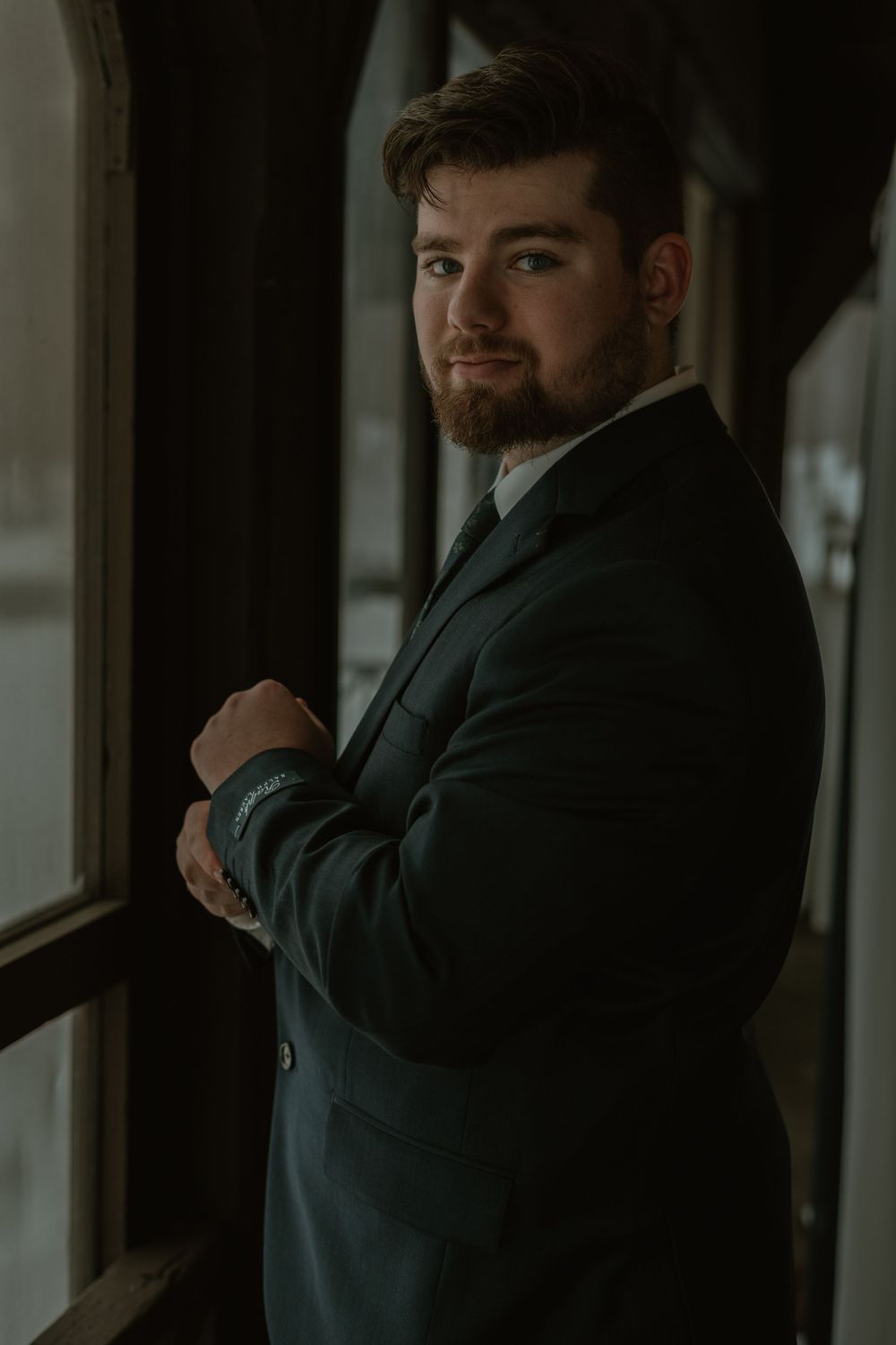 Dapper groom in dark suit posing by window with arms crossed in moody indoor portrait.
