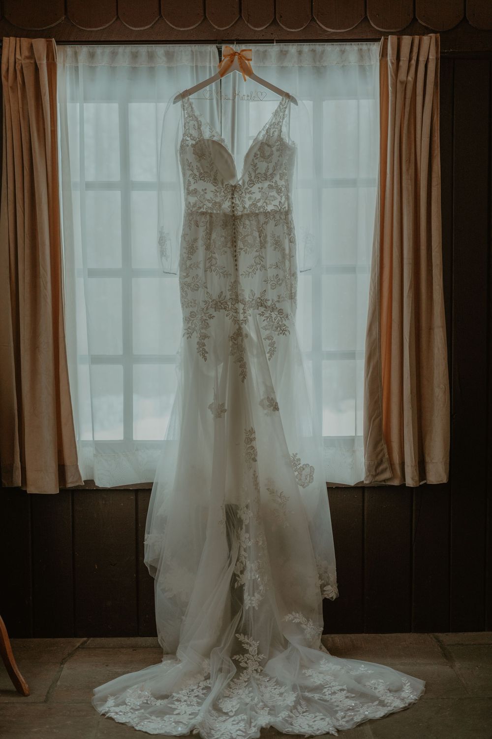 Elegant lace wedding gown with long train hanging on hanger in front of curtained window.